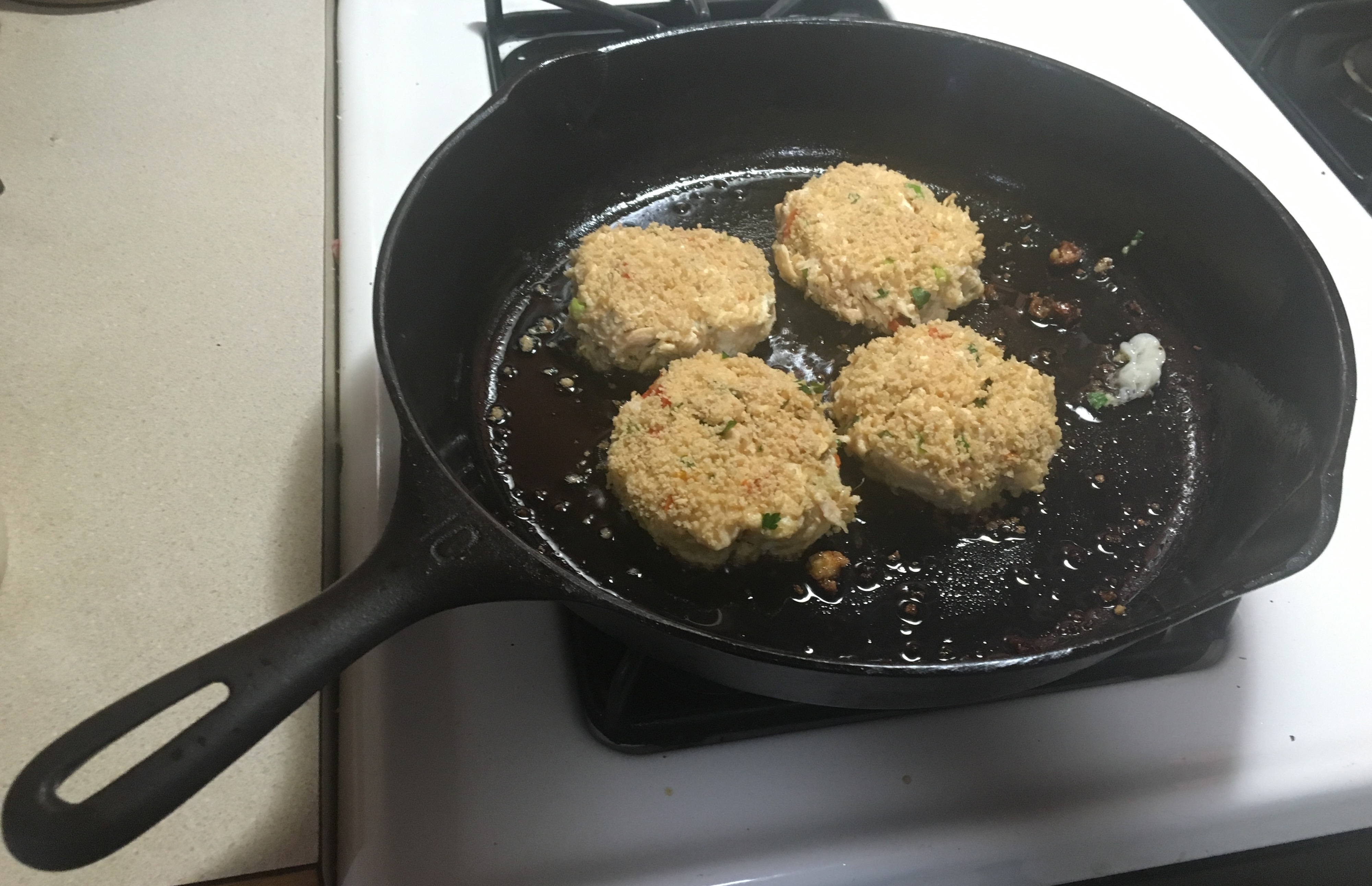 Four breaded lake trout cakes cooking in a cast-iron skillet